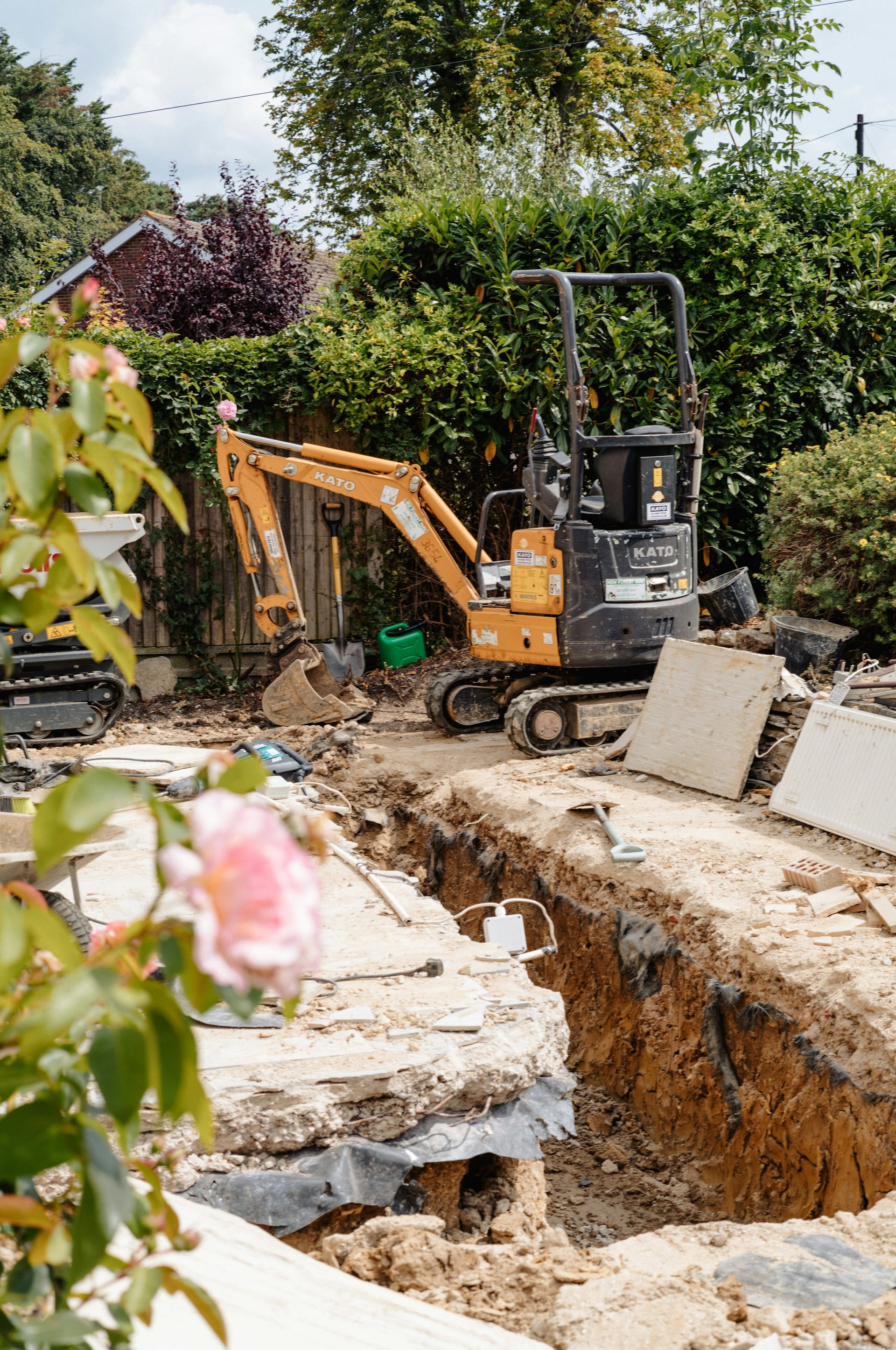 A mini excavator parked beside an open footing trench in a hedged back garden